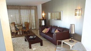 A living room with a brown sofa, a coffee table, a side table with a lamp, a patterned rug, and a dining area with a table and chairs. - Luxury apartment in private condominium, with pool (Quarteira)