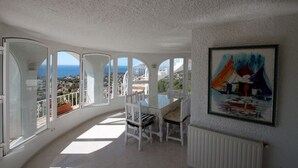 Una stanza luminosa con un tavolo e sedie in vetro, un quadro appeso al muro e vista sul mare e sulla città attraverso ampie finestre.