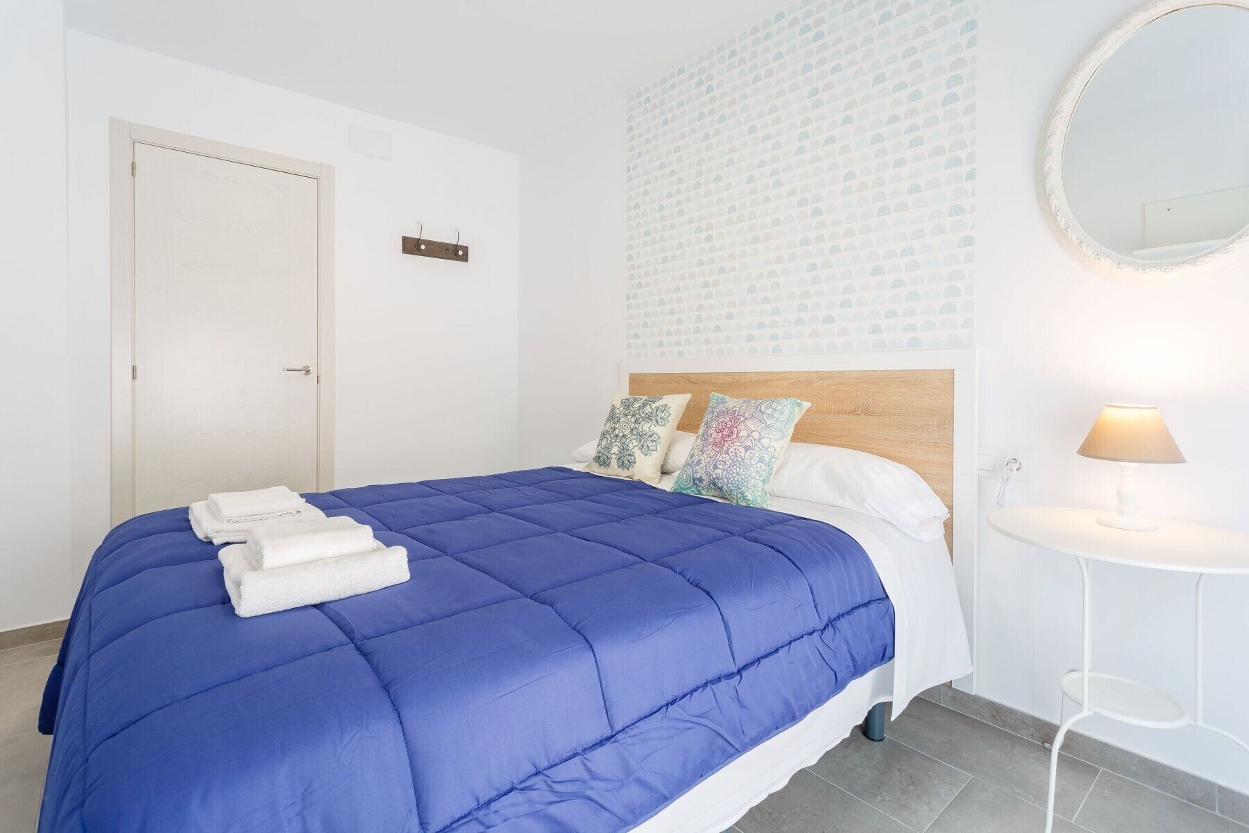 A neatly made bed with a blue quilt, white pillows, and a small bedside table with a lamp.