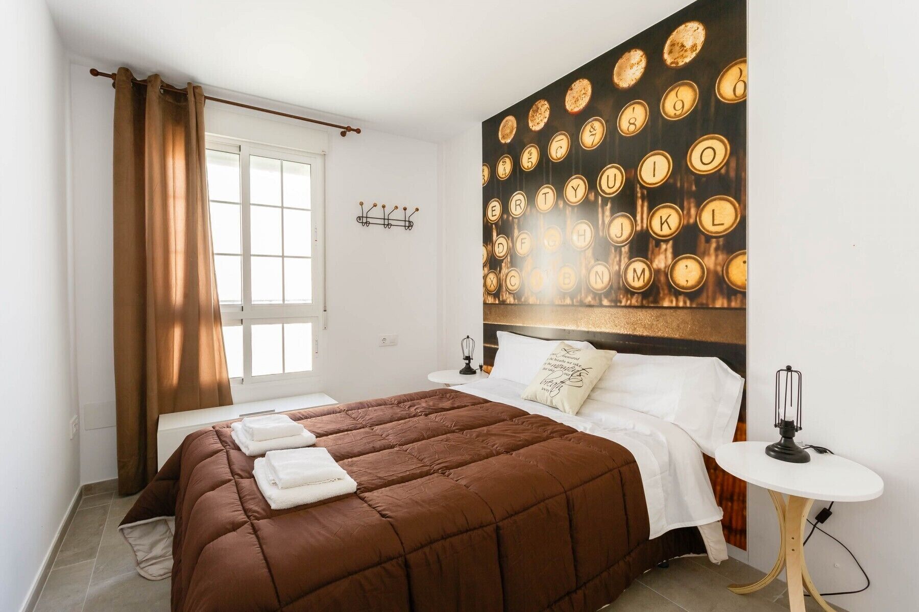 Ein Schlafzimmer mit einer braunen gesteppten Bettdecke, weißer Bettwäsche, einer wandmontierten Leuchte mit runden Glühbirnen, einem kleinen Nachttisch mit Lampe und einem Fenster mit Vorhängen.