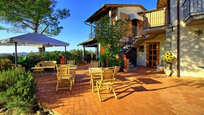 Eine Terrasse mit Korbmöbeln und einem überdachten Bereich, ein zweistöckiges Gebäude mit Balkon sowie eine Landschaftsaufnahme.