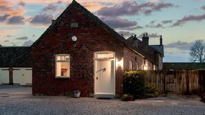A small brick house with a white door and windows, a gravel driveway, and a wooden fence in the background. - The Stables - at Broadgate Farm Cottages (Beverley)