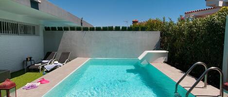 Une piscine rectangulaire à l’eau d’un bleu limpide, entourée d’une terrasse carrelée. On y trouve des chaises longues et une petite table agrémentée d’une plante.