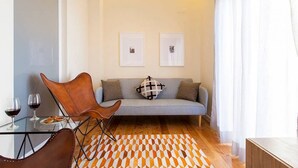 Een moderne woonkamer met een bank, twee ingelijste foto’s aan de muur, een glazen tafel met wijnglazen en een bord eten, een tapijt met een patroon, en een lederen fauteuil.