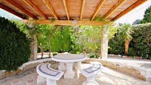 A stone patio with a round table and two stone benches under a wooden pergola. - Kathrin - beautiful little house in pretty grounds with lovely views in Benissa (Benissa)