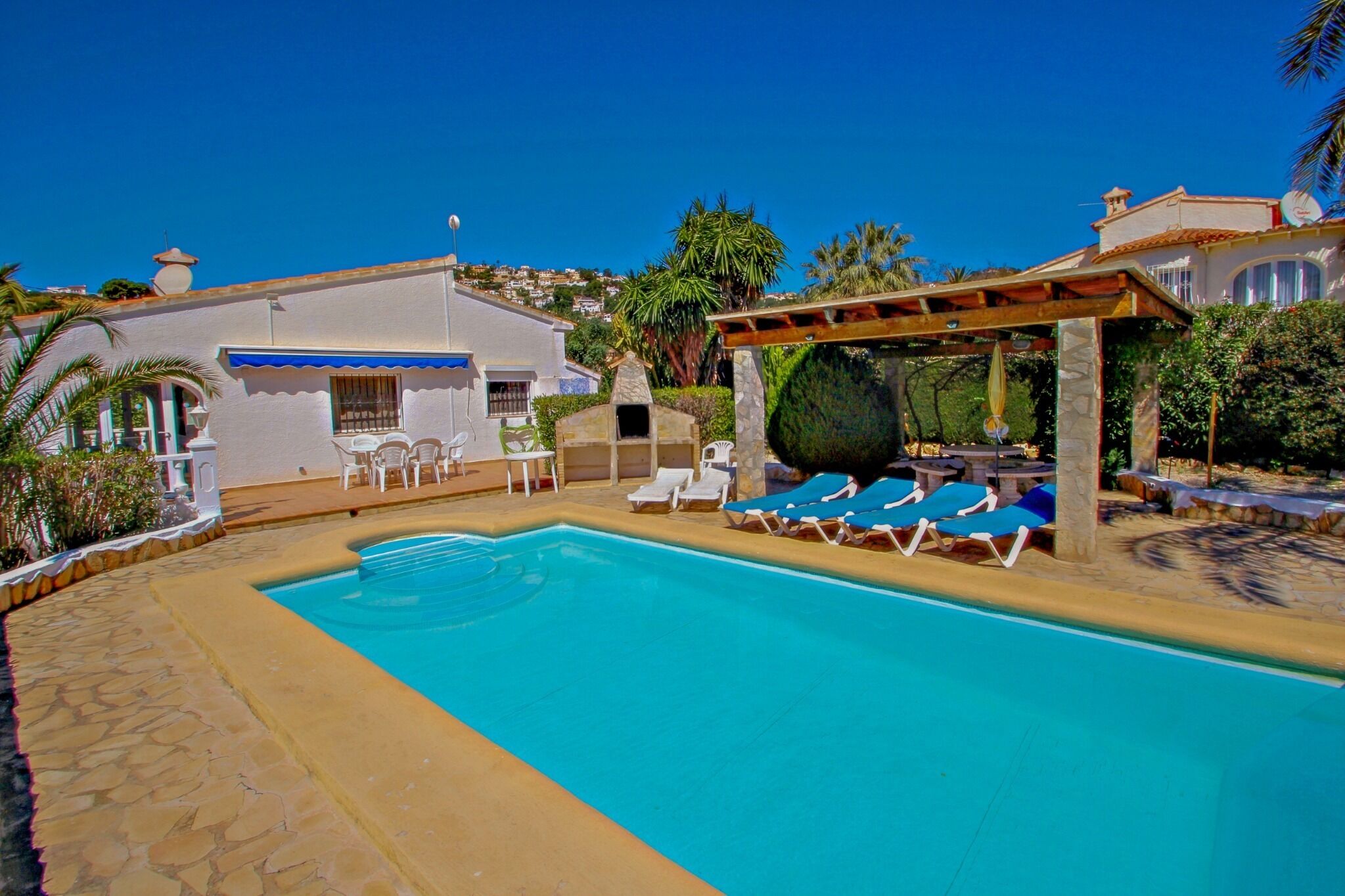 Une piscine avec des chaises longues, une pergola et un petit bâtiment surmonté d’une marquise bleue.
