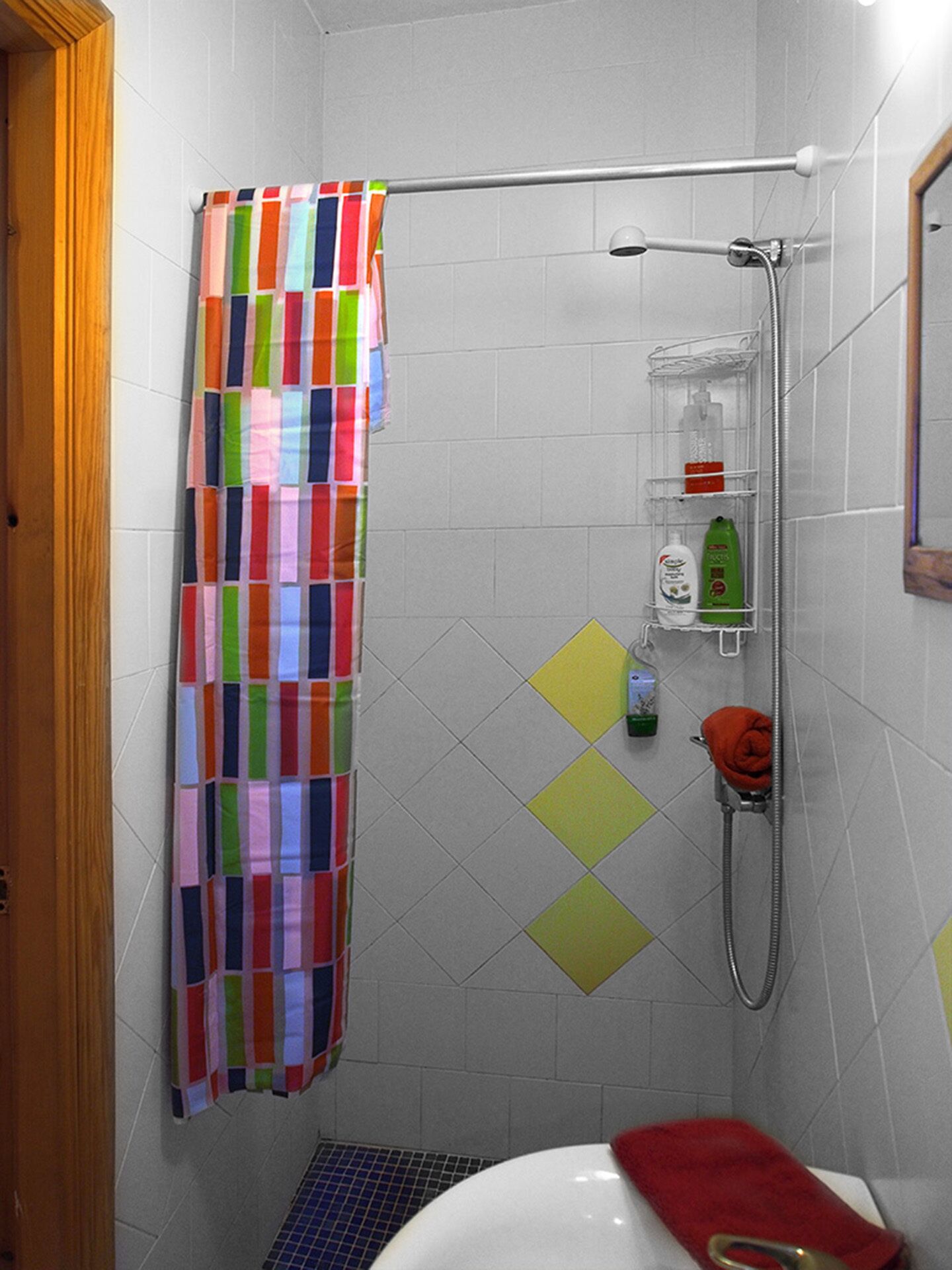 A small bathroom with a shower, a toilet, and a sink. There is a colorful shower curtain, a towel rack, and a shelf with toiletries.