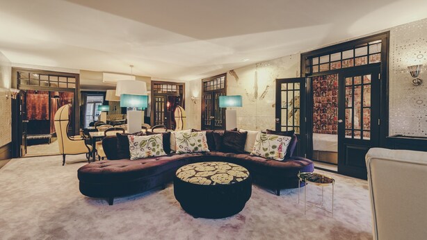 A living room with a sectional sofa, a round ottoman, and a small side table. There is a door with a decorative panel and a wall-mounted lamp. - Master Deco Gem in Bica (Lisboa)
