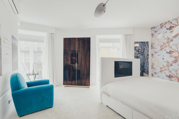 A modern bedroom with a large bed, a blue armchair, a wooden wardrobe, and a TV on the wall.