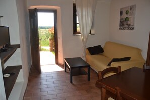 Un salon avec un canapé, une petite table et un téléviseur posé sur un meuble. Il y a une fenêtre avec des rideaux et une vue sur la verdure à l’extérieur.