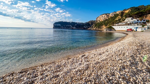 A pebble beach with clear water, a rocky cliff, and buildings on the hillside. - Casa Luna - Javea, Costa Blanca (Javea)