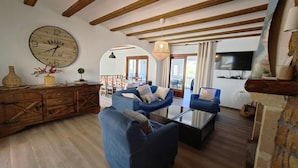 A living room with a large clock, blue sofas, wooden beams, and a dining area visible through an archway. - Casa Luna - Javea, Costa Blanca (Javea)