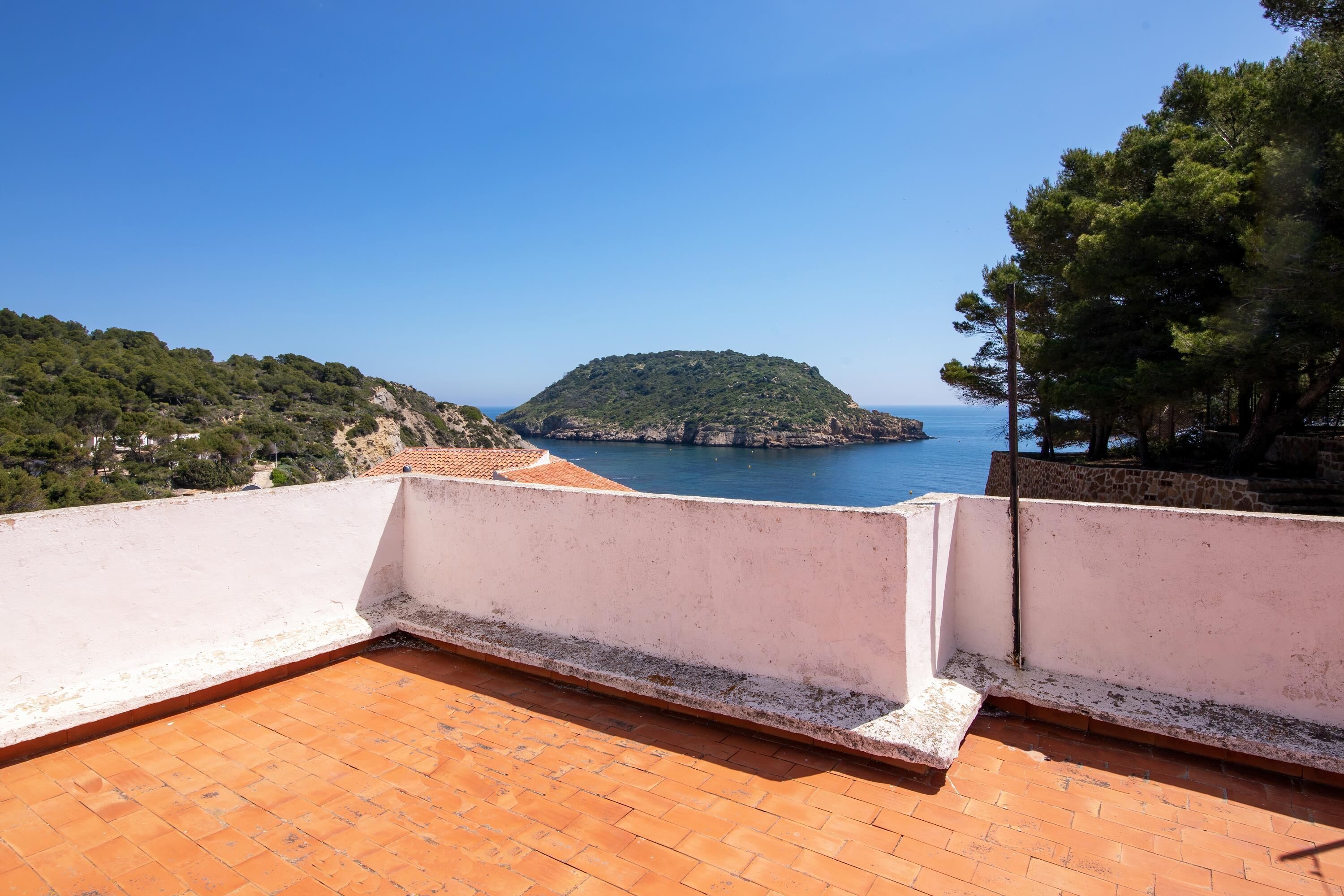 A rooftop terrace with terracotta tiles, overlooking a coastal landscape with a hill and the sea.