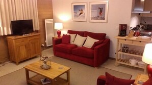 A living room with a red sofa, wooden coffee table, and a television on a wooden cabinet. There are two framed pictures on the wall, a lamp on the side table, and a shelf with kitchen items.