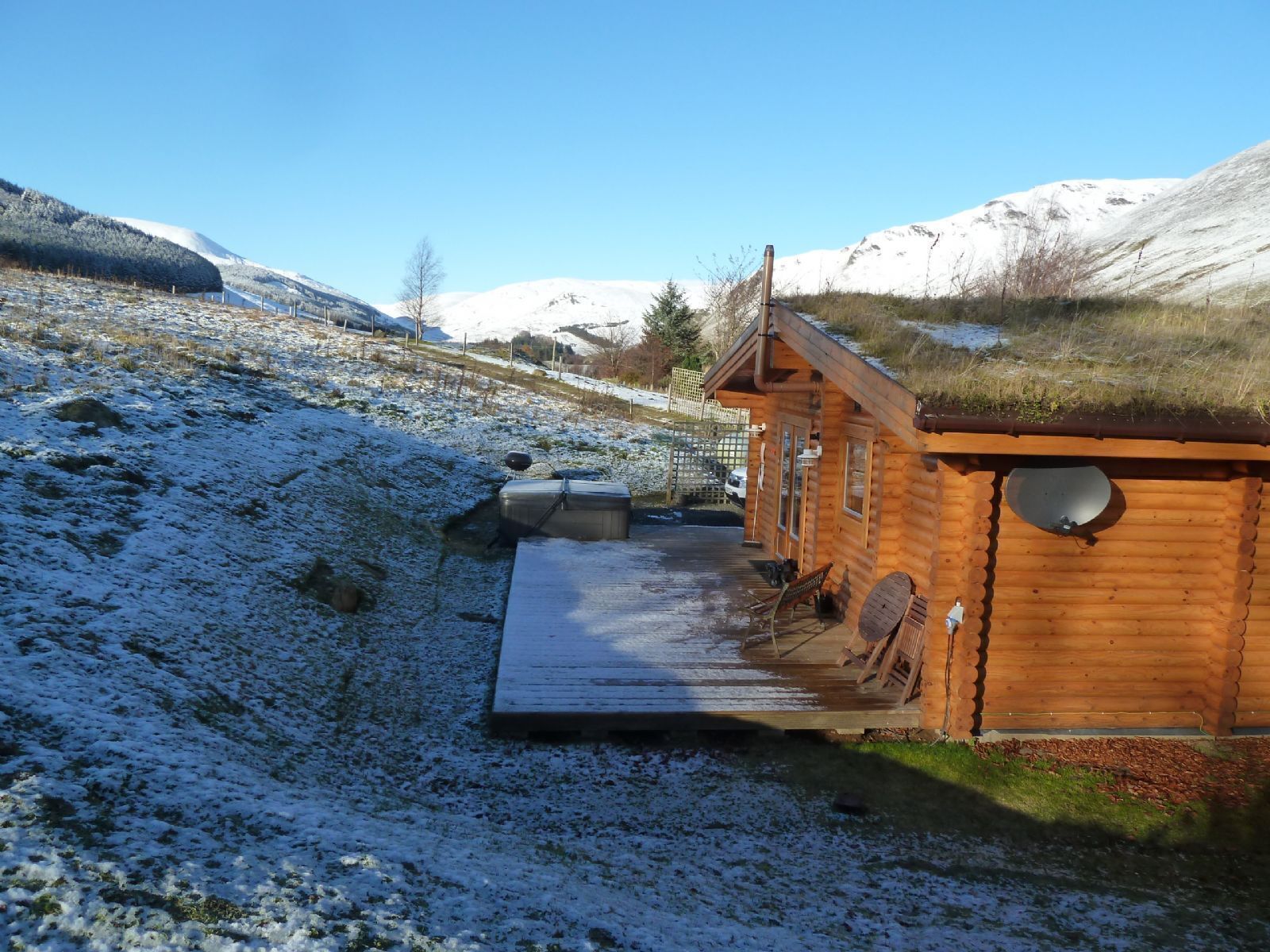 A wooden cabin with a deck, surrounded by snow-covered hills and clear skies.
