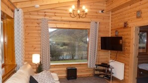 Ein gemütliches Zimmer mit Holzvertäfelung, einer Couch, einem Fernseher und einem großen Fenster mit Blick auf einen See und Berge.