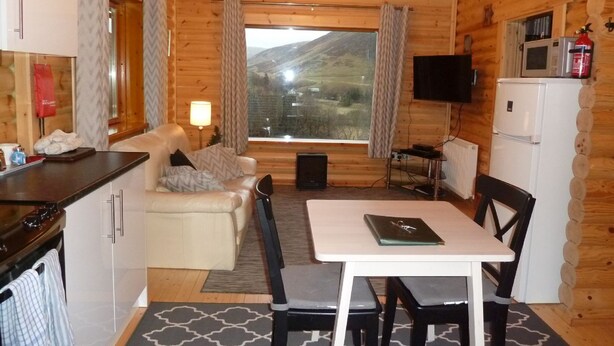 Ein gemütliches Blockhaus mit einer Couch, einem Esstisch, Stühlen, einem Fernseher und einer Küchenzeile.