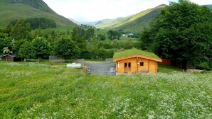Eine rustikale Holzhütte mit grünem Dach in einem üppigen Tal, umgeben von Wildblumen und Bäumen.