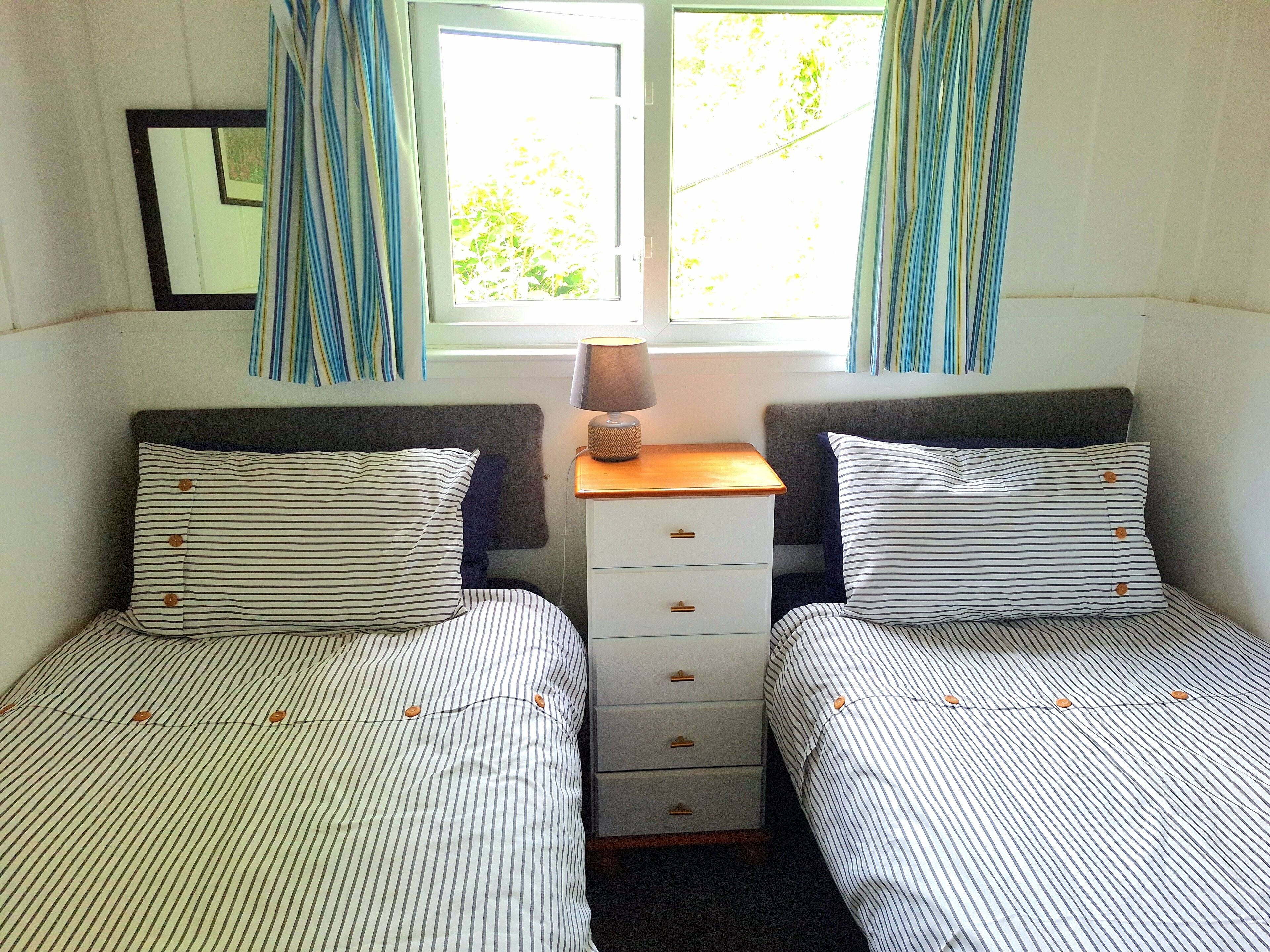 A bedroom with two beds, each with striped bedding and matching pillows, a bedside table with a lamp, and a window with curtains.