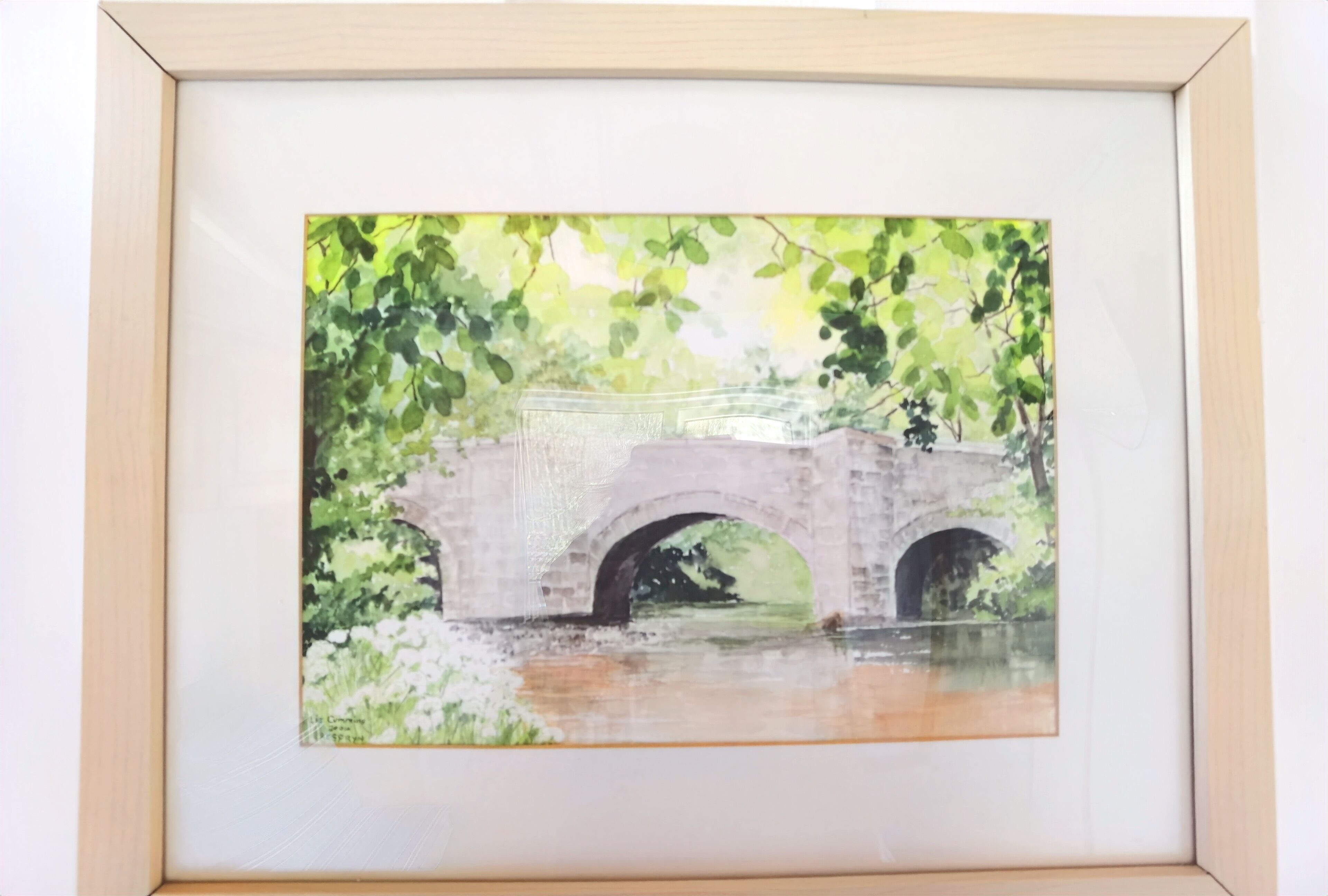Un quadro incorniciato raffigura un ponte in pietra con archi, circondato dal verde.