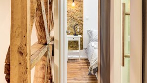 Un pasillo estrecho con una rústica escalera de madera a la izquierda, que lleva a un dormitorio con pared de piedra y una lámpara de noche.