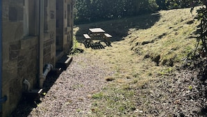 Un bâtiment en pierre doté d’une cheminée, un chemin de gravier et une table de pique-nique, le tout situé dans un espace verdoyant bordé d’arbres.