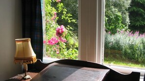 Une pièce avec une table en bois, une lampe et une vue sur un jardin où poussent des fleurs roses, visible à travers de grandes fenêtres.