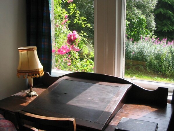 Ein Zimmer mit einem Holztisch, einer Lampe und Blick auf einen Garten mit rosa Blumen durch große Fenster.