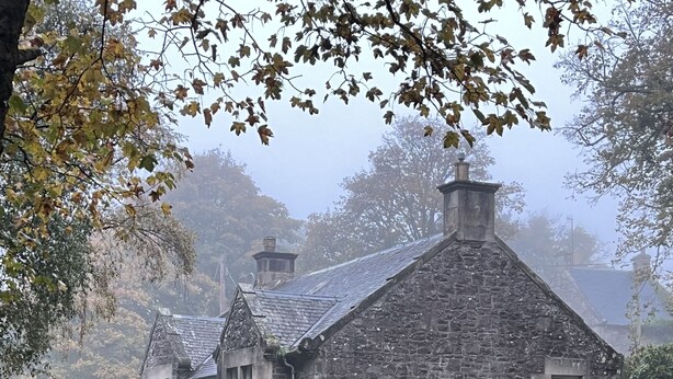 Un bâtiment en pierre à l’architecture traditionnelle, une pelouse verdoyante et un sentier jonché de feuilles mortes.