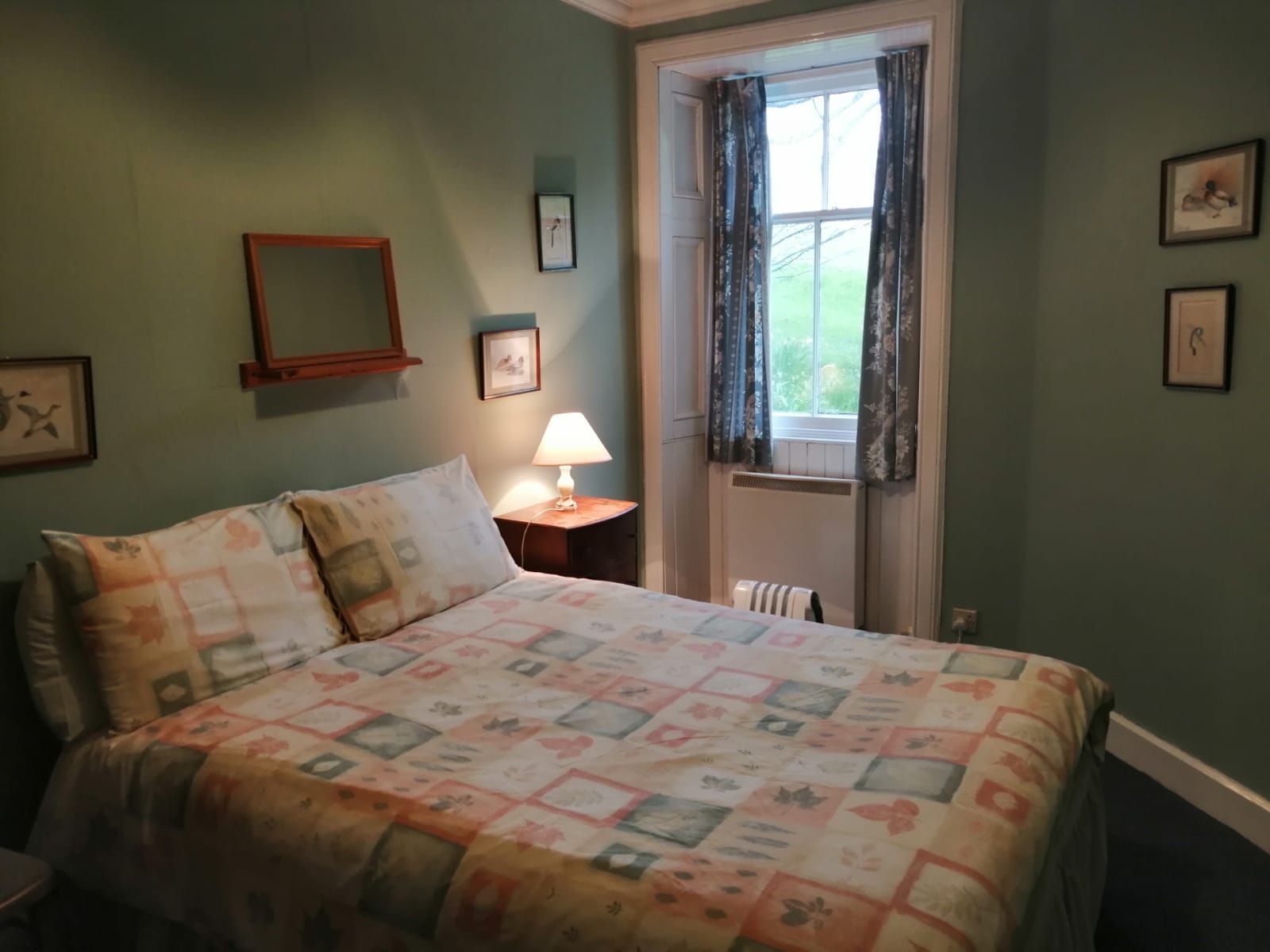 A bedroom with a bed, a nightstand, a lamp, a window with curtains, and framed pictures on the wall.