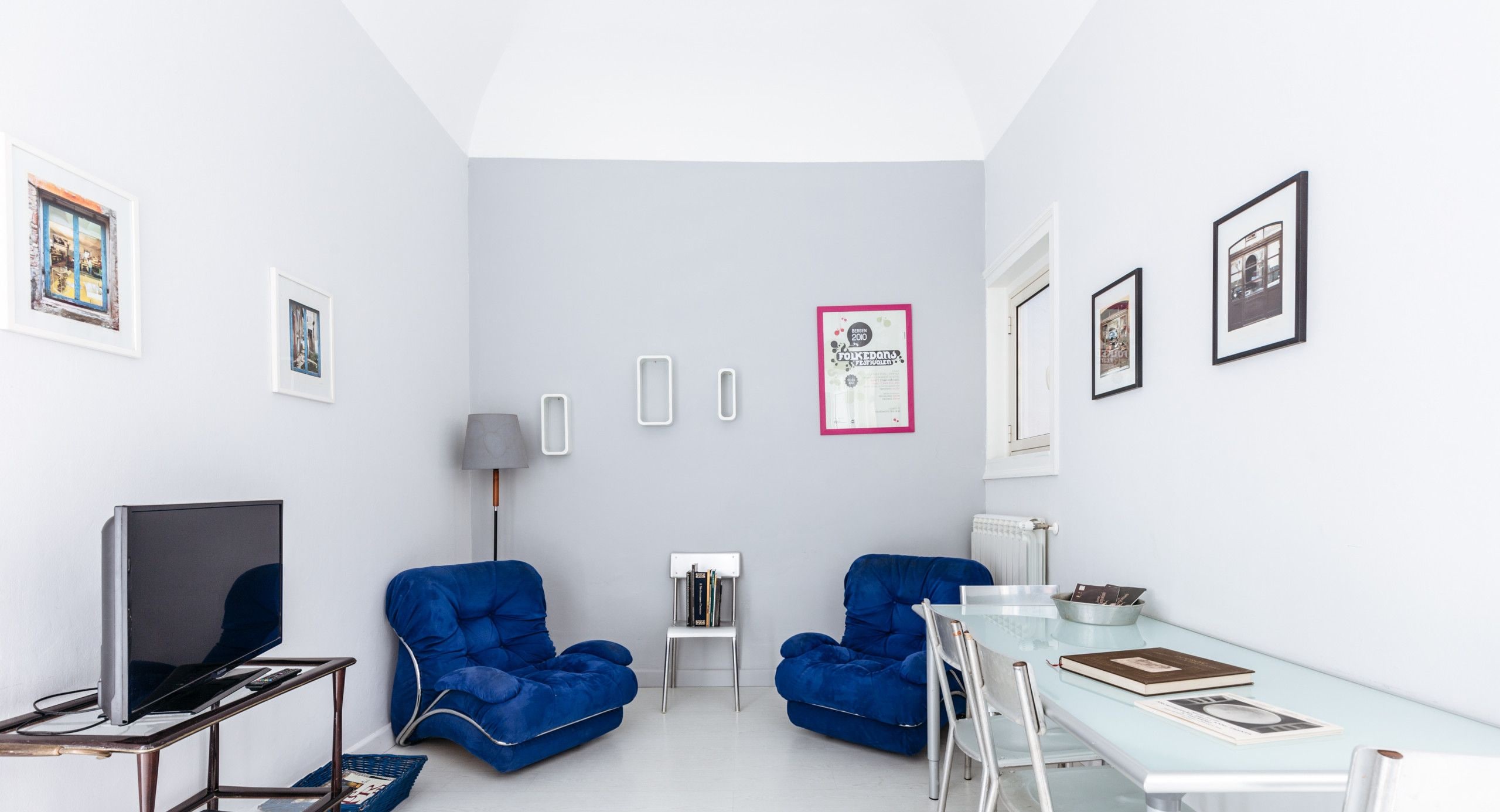 A modern, well-lit room with a white floor, a glass dining table, two blue armchairs, a small white side table, a TV on a metal stand, and framed pictures on the walls.