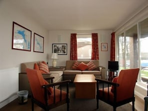 En stue med sofa, to lænestole, sofabord og fjernsyn. På væggen hænger billeder i rammer, og på et sofabord står en lampe. Der er et vindue med gardiner.