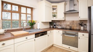 A modern kitchen with white cabinetry, a stainless steel range hood, and a window with blinds. - Teagles Cottage - a delightful home from home in Stow-on-the-Wold (Cheltenham)