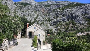 Una casa de piedra con un pequeño porche y una reja, rodeada de colinas rocosas y vegetación.