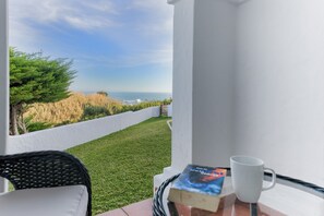 Een balkon met uitzicht op de oceaan, een witte muur, een rieten stoel, een tafel met een boek en een witte mok.