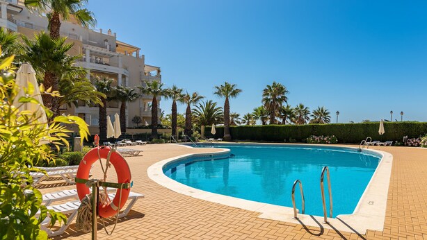 A swimming pool with lounge chairs and a lifebuoy, surrounded by palm trees and a building with balconies. - Las Dunas - Punta del Moral Beach PLHD115P113 (Punta del Moral)