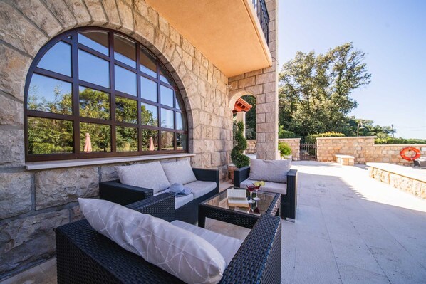A stone building with a large arched window, outdoor seating with wicker furniture, and a view of trees.
