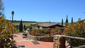 A stone pathway leading to a rustic building with a tiled roof, surrounded by lush greenery and a clear blue sky.