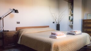 Een opgemaakt bed met een beige sprei, een bedlamp, een vaas met takken, en een boekenplank vol boeken.
