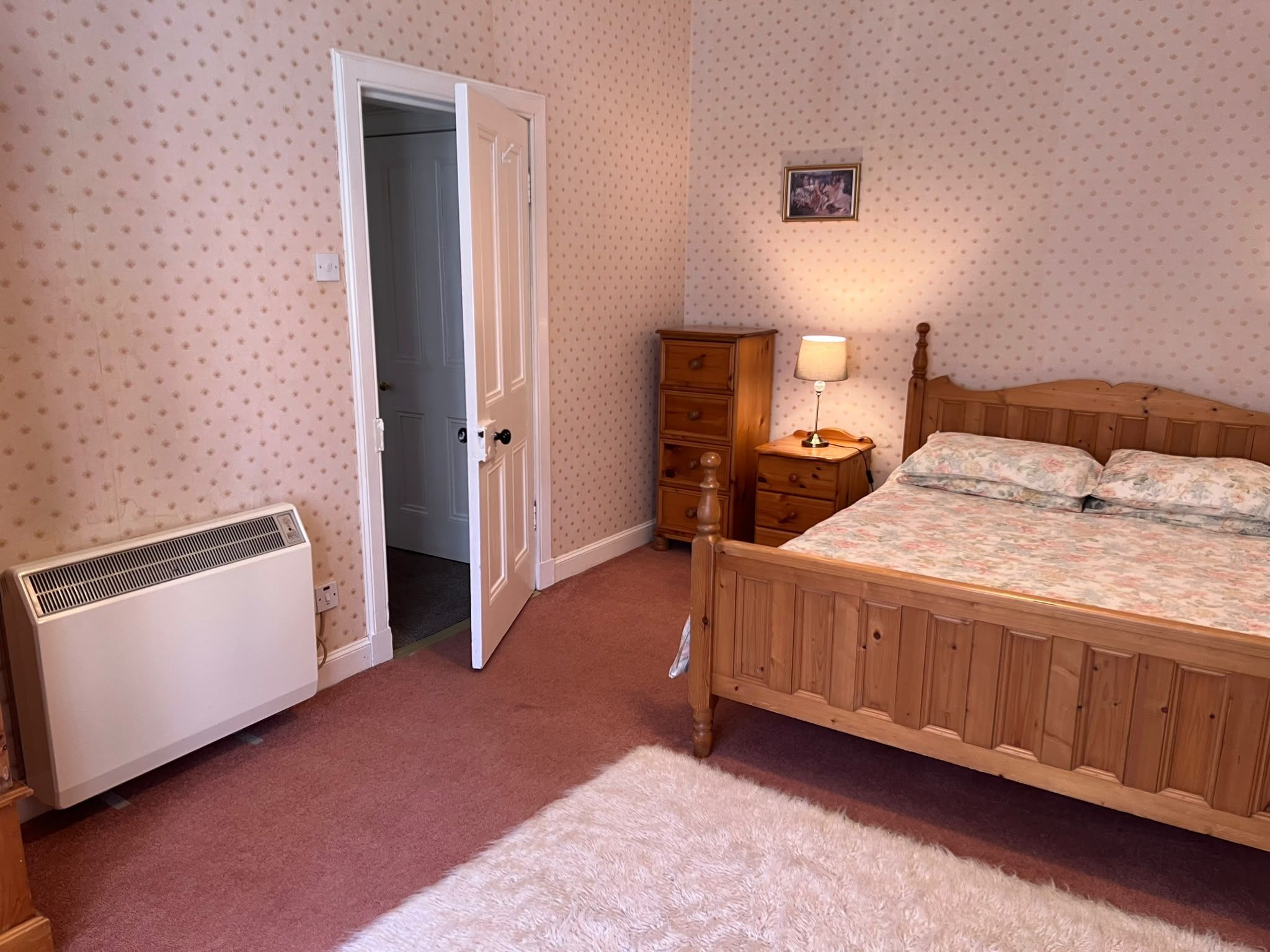A bedroom with a wooden bed, a nightstand with a lamp, a dresser, a radiator, and a door leading to another room.