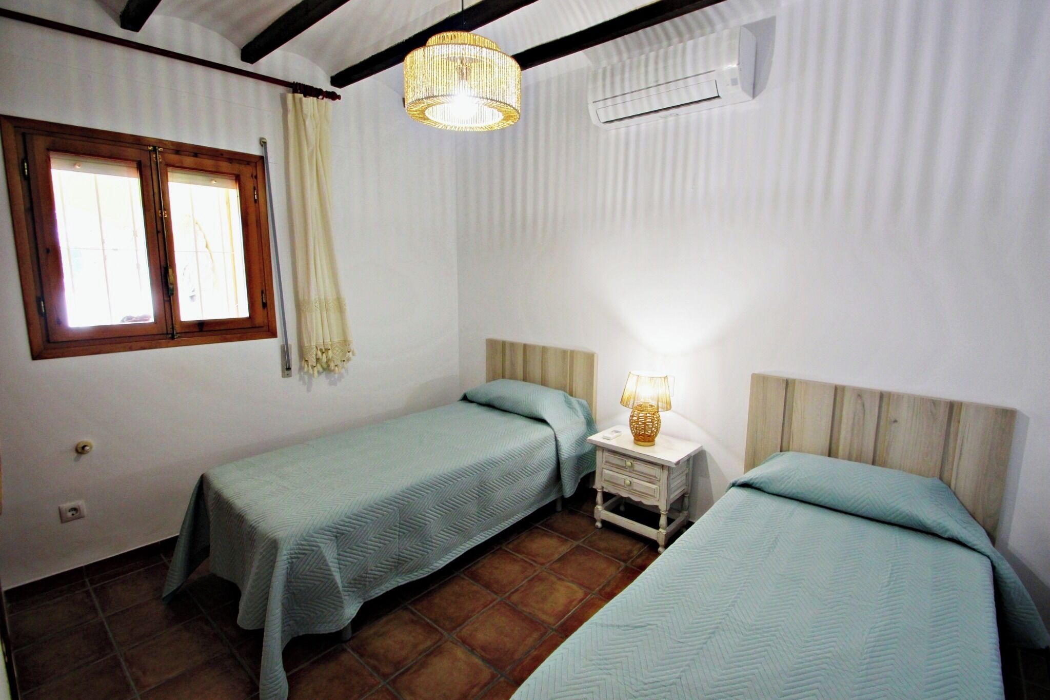 Une chambre avec deux lits, une petite table de chevet avec une lampe, une fenêtre avec des rideaux et un appareil de climatisation.