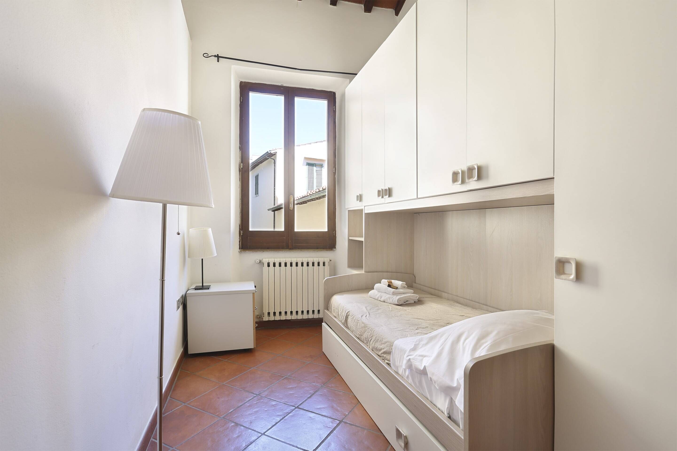 Une petite chambre bien éclairée, avec un lit simple, une table de chevet, une lampe et une fenêtre.
