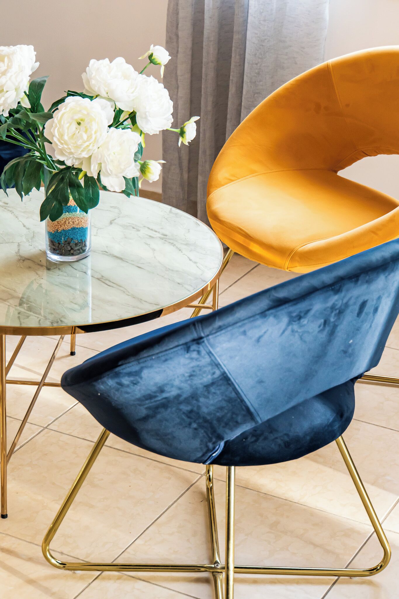 A dining area with a round glass table, a yellow upholstered chair, a blue upholstered chair, and a vase with white flowers.