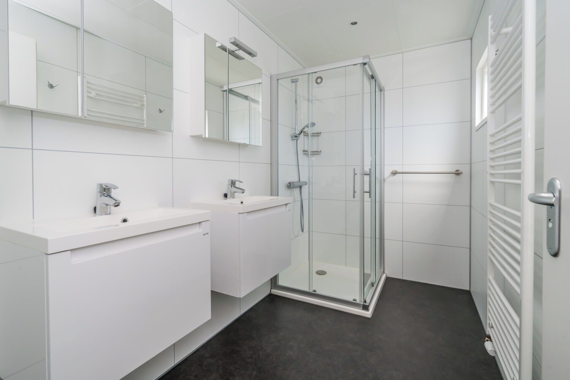 Un baño moderno con dos lavamanos, una cabina de ducha de vidrio y suelo de baldosas blancas.