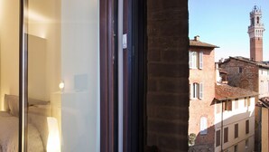 A hotel room with a bed, a nightstand, and a window overlooking a historic building with a tower. - THE TOLOMEI - apartment in the heart of the main street (Siena)