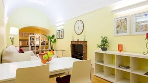 A living room with a white sofa, a dining table with a fruit bowl, a fireplace, and a bookshelf.