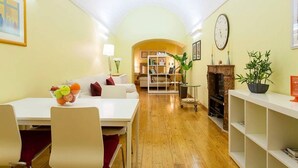 A cozy living room with a white sofa, a dining table with chairs, a bookshelf, a fireplace, and a clock on the wall.