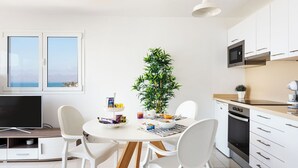 A modern kitchen with white cabinets, a dining table with chairs, a TV, and a large window with a view of the sea. - ESTELAI by Buenavilla | Apartment with breathtaking front line Ocean views (Tías)