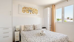A bedroom with a bed, a nightstand, a dresser, a painting of a tree, and a window with curtains. - ESTELAI by Buenavilla | Apartment with breathtaking front line Ocean views (Tías)