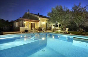 Une piscine avec des chaises longues et un espace patio devant une maison, de nuit.